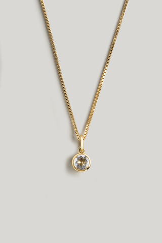A gold chain necklace with a small round bezel-set clear gemstone pendant, displayed against a solid light gray background.