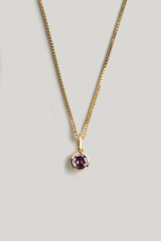 A gold chain necklace with a small bezel-set oval purple gemstone pendant, displayed against a neutral light gray background.