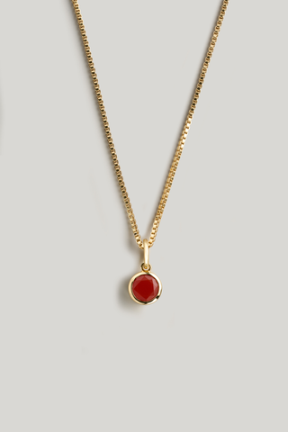 A gold chain necklace with a small oval red gemstone pendant, displayed against a neutral light gray background.