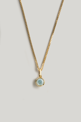 A gold chain necklace with a small round bezel pendant containing a light blue-green stone, displayed against a neutral light gray background.