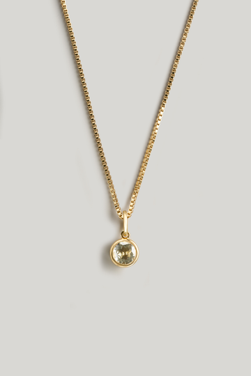 A gold chain necklace with a small round bezel-set pale yellow-green gemstone pendant, displayed against a neutral light gray background.