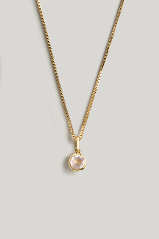 A delicate gold chain necklace with a small round bezel-set pale pink stone pendant, displayed against a solid light gray background.