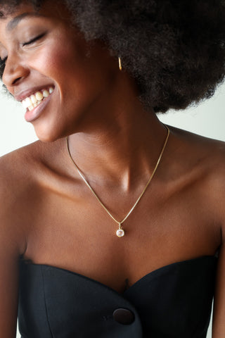 A close-up lifestyle shot of a smiling woman with dark curly hair wearing a delicate gold chain necklace with a small round pendant featuring a dark blue stone, paired with a black strapless top.
