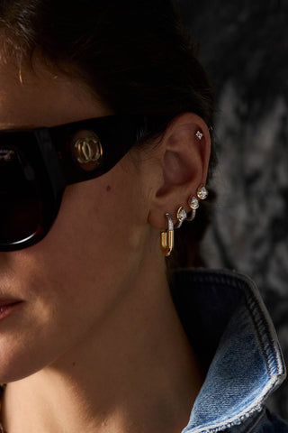 A close-up of a person's ear wearing multiple gold and silver hoop and stud earrings, with dark sunglasses and a denim jacket visible in a dimly lit setting.