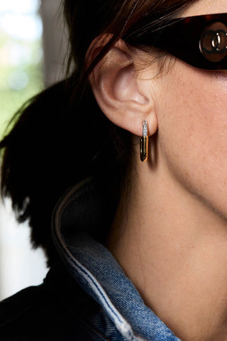 A close-up of a person's ear wearing a small gold hoop earring with a vertical row of clear stones, paired with dark sunglasses and a denim jacket.
