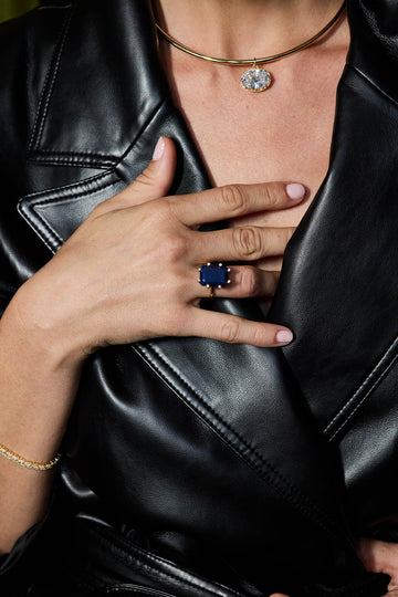 A close-up lifestyle shot of a person wearing a black leather jacket, a gold chain necklace with a clear stone pendant, a gold bracelet, and a ring with a large rectangular dark blue gemstone.
