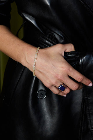 Roxy tennis bracelet, gold plated, worn on the wrist with a deep blue rectangular stone ring and a black leather coat
