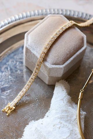 Roxy tennis bracelet, gold plated, draped over a cream velvet ring box on a silver-tone tray with lace
