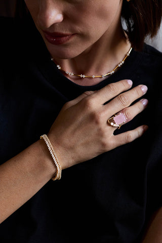 Gold plated Nico and Clara tennis bracelets on the wrist, hand on a black top with a pink stone ring and a station necklace