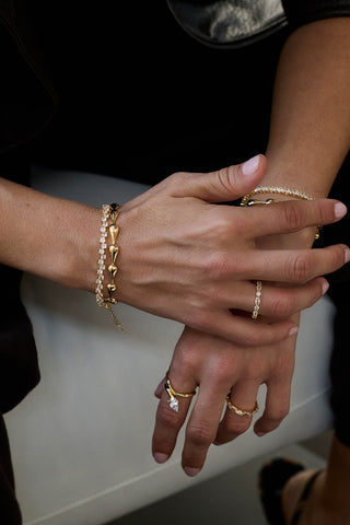 Gold plated Riva bracelet stacked with a tennis bracelet on crossed hands, with a marquise stone ring and stacked bands