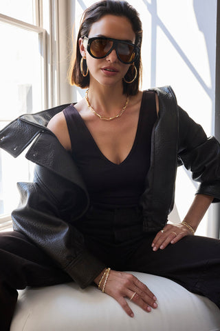 Model in sunglasses and a leather jacket seated by a window wearing gold-tone tennis bracelets, rings and a paperclip chain
