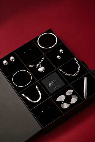 A black jewelry gift box with multiple compartments displaying silver-toned jewelry, including earrings, rings, a pendant, chain links, and a cylindrical piece, set against a deep red background.