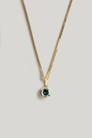 A gold chain necklace with a small round bezel-set dark blue gemstone pendant, displayed against a neutral light gray background.